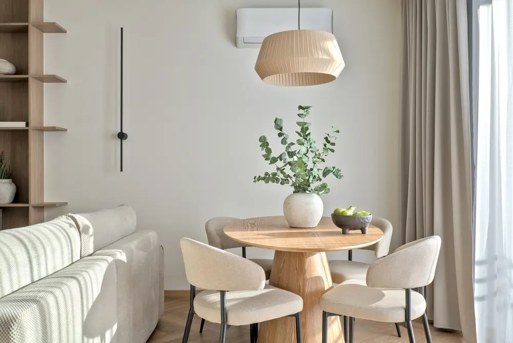 Coin repas chaleureux avec une table ronde en bois et quatre chaises beiges. Un grand vase de verdure et une coupe de pommes ornent la table. Un éclairage tamisé crée une ambiance sereine.