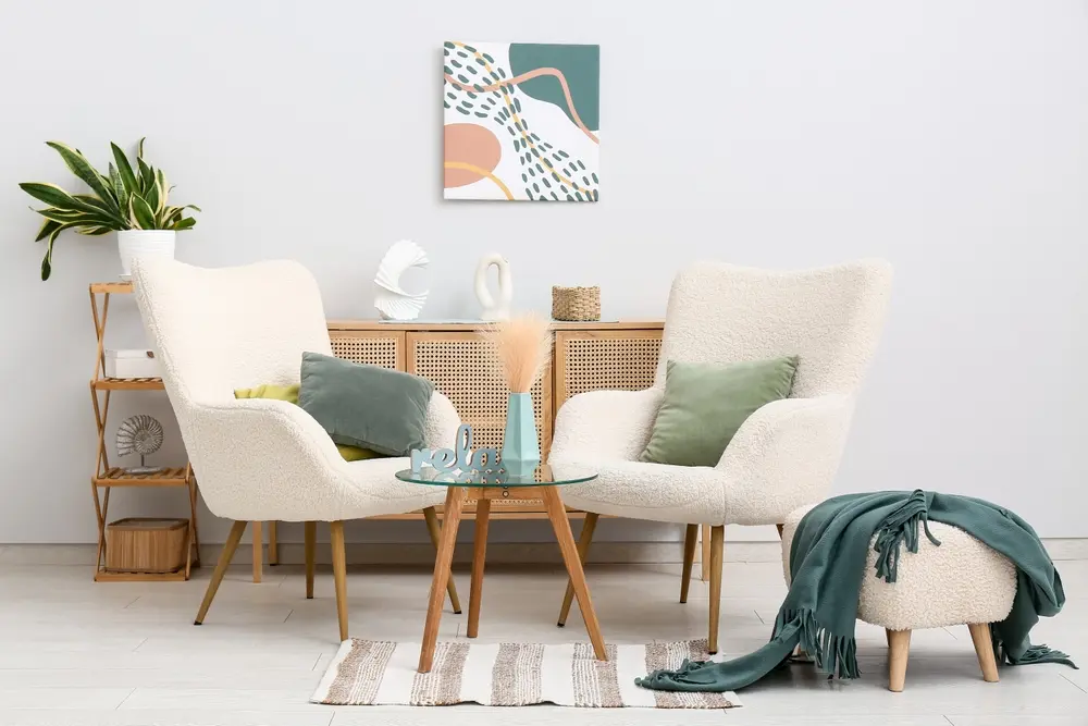 Un coin salon chaleureux avec deux fauteuils crème aux coussins verts, une table basse en verre et un petit pouf. Un buffet orné d'objets décoratifs, une plante et une œuvre d'art abstraite créent une ambiance sereine et moderne.