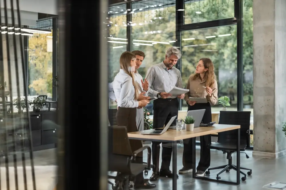 Quatre personnes en tenue de ville discutent avec enthousiasme de documents dans un bureau moderne baigné de lumière naturelle, grâce à de grandes fenêtres et un ordinateur portable posé sur la table.
