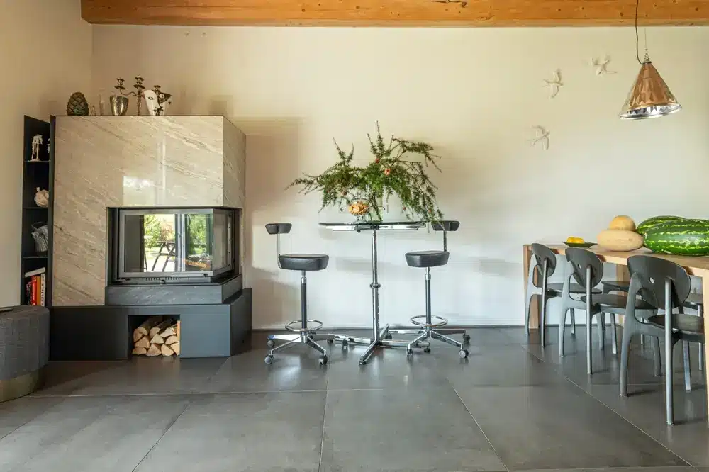 Cuisine moderne avec une grande cheminée en marbre sur la gauche, une table haute avec tabourets et un centre de table végétal au centre, et une table à manger avec des fruits.