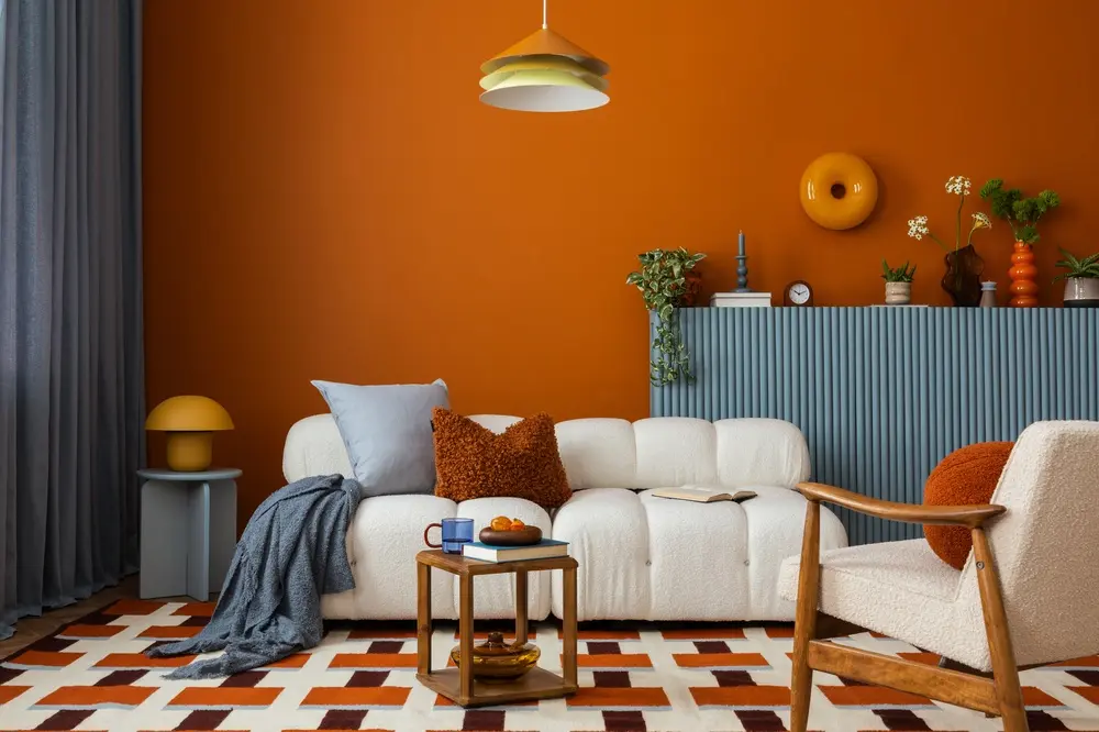Un salon chaleureux avec un mur d'accent orange, un canapé blanc et un tapis à motifs. La décoration comprend des coussins, des plantes et une petite table avec une tasse et un bol.