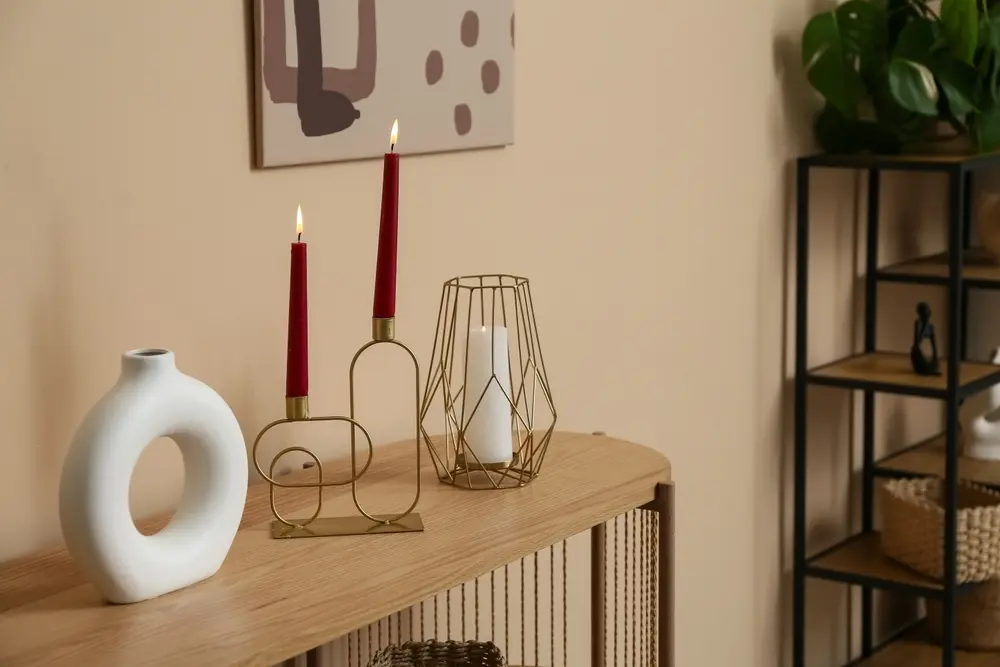 Une table en bois minimaliste avec des bougies rouges dans des bougeoirs géométriques, un vase rond et une lanterne en fil de fer. Des tons neutres et des œuvres d'art créent une ambiance chaleureuse.