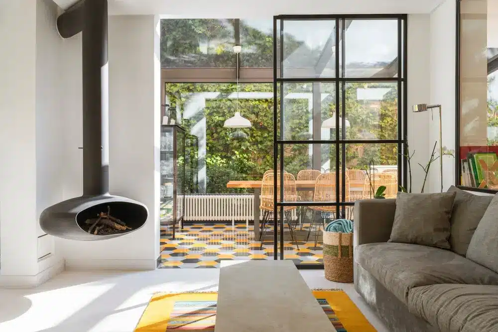 Salon moderne avec un canapé gris confortable et une cheminée suspendue. Des baies vitrées laissent entrevoir une salle à manger baignée de lumière, ornée de carreaux à motifs et de chaises en rotin, créant une ambiance chaleureuse et accueillante.