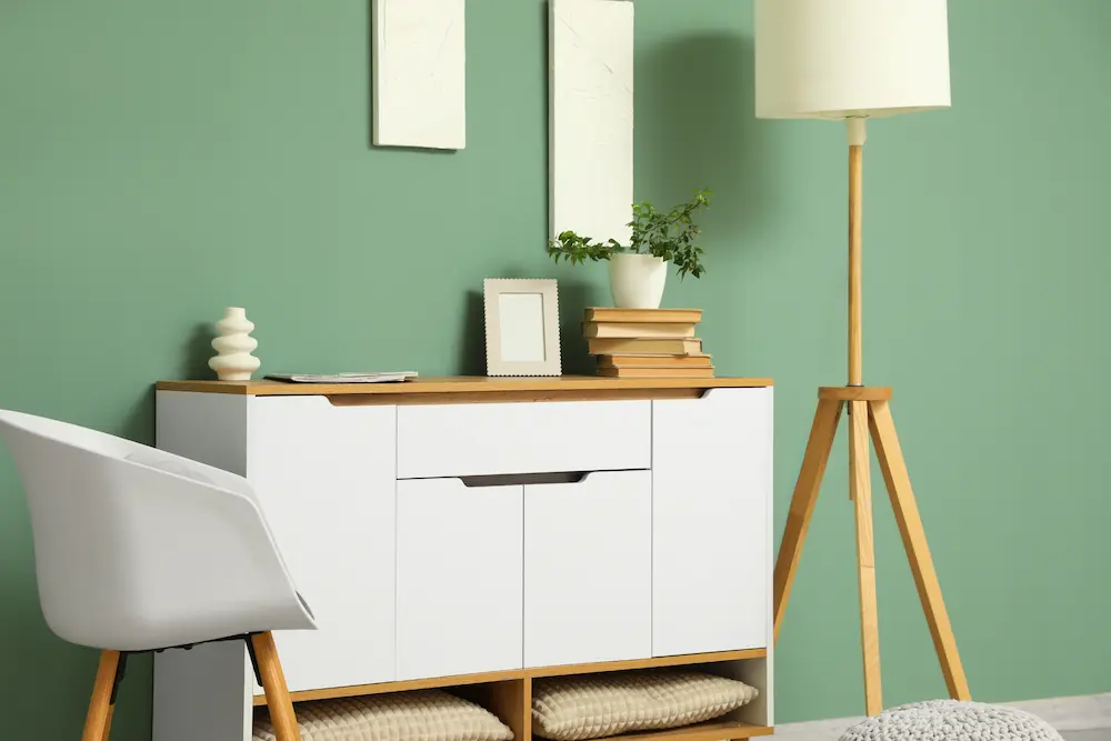 Chambre minimaliste avec un mur vert, une armoire blanche et des touches de bois. La décoration comprend une plante en pot, une pile de livres, une photo encadrée et une lampe sur pied.