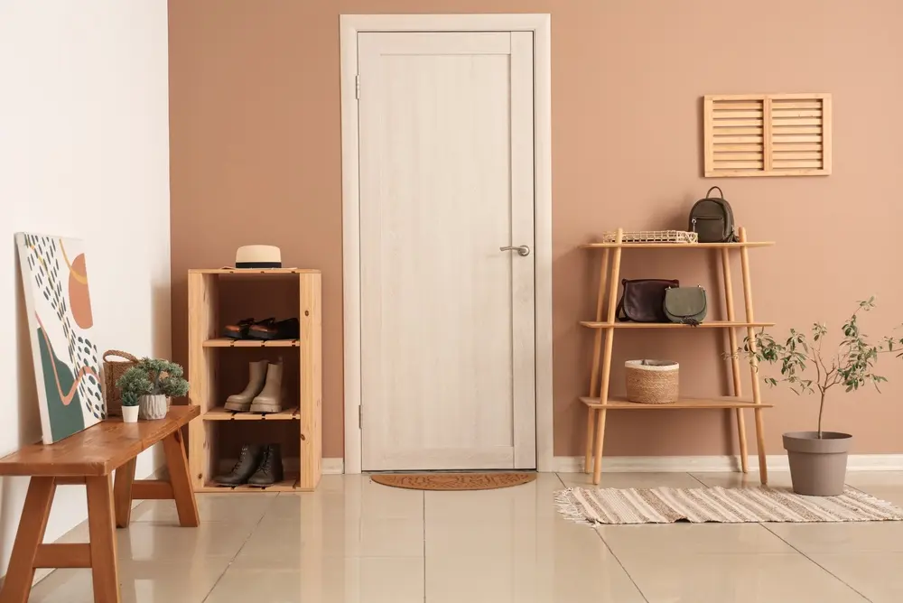 Entrée minimaliste avec porte en bois clair, murs beiges et sol carrelé. Des étagères en bois accueillent chaussures, sacs et objets de décoration. Une plante en pot apporte une touche de verdure.