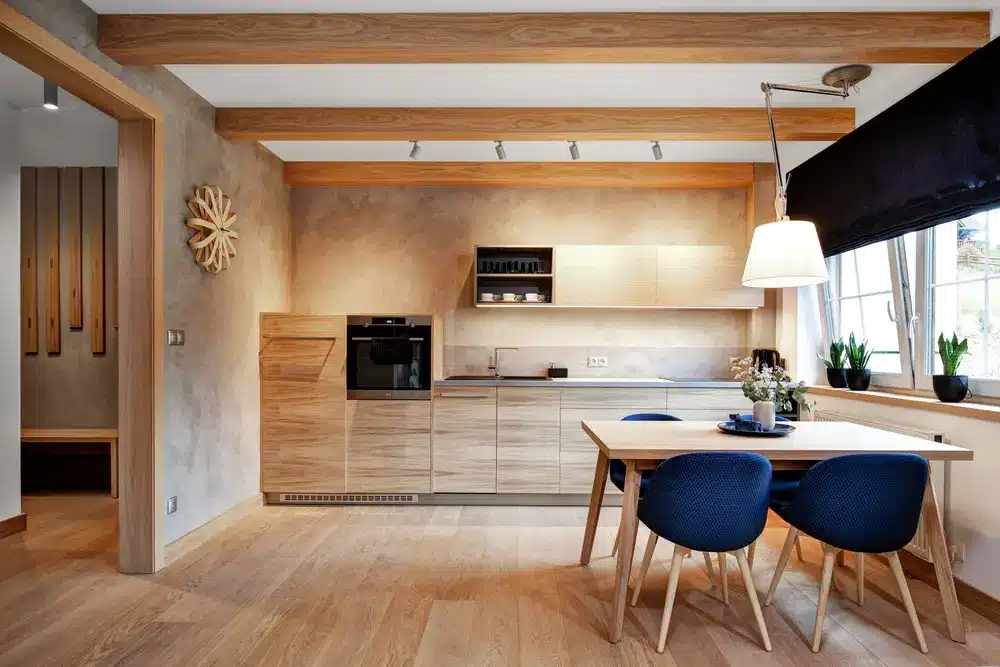 Cuisine moderne avec poutres apparentes, placards en bois clair et table à manger entourée de chaises bleues. La pièce est baignée d'une douce lumière grâce aux plantes qui éclairent la pièce depuis une grande fenêtre.