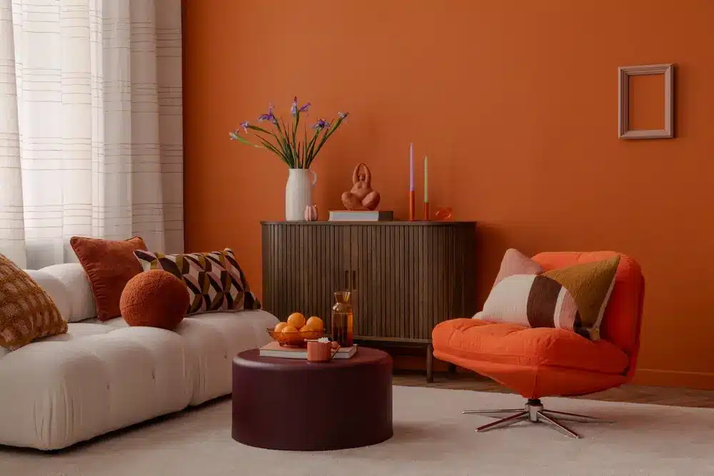 Salon chaleureux avec un mur d'accent orange, un canapé blanc et un fauteuil orange. Une table ronde accueille des fruits et des bougies. Un vase de fleurs orne une armoire.