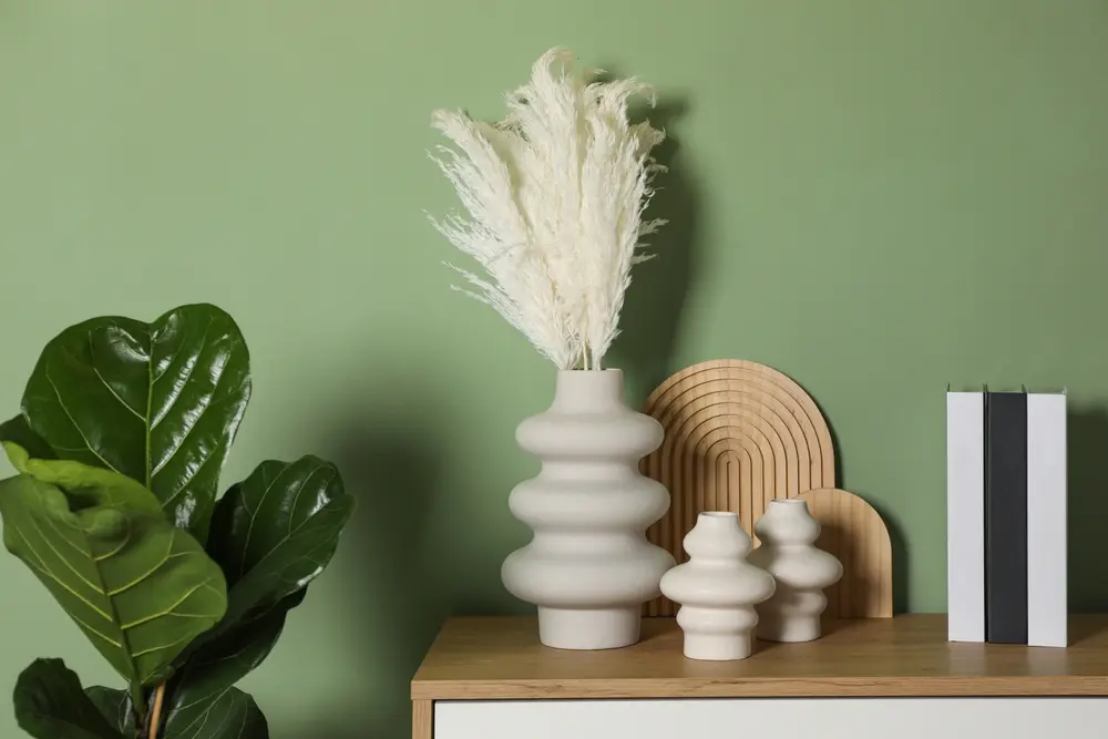 Des vases abstraits remplis d'herbes de la pampa sont posés sur une étagère en bois contre un mur vert, accompagnés d'un figuier lyre et de livres décoratifs, créant une atmosphère sereine.