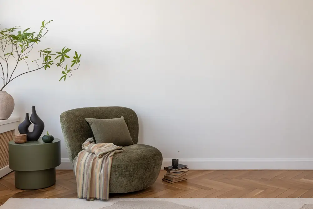 Dans cette pièce chaleureuse, un fauteuil vert olive agrémenté d'un coussin et d'un plaid est placé à côté d'une petite table ronde ornée de vases. Une plante en pot apporte une touche de fraîcheur.