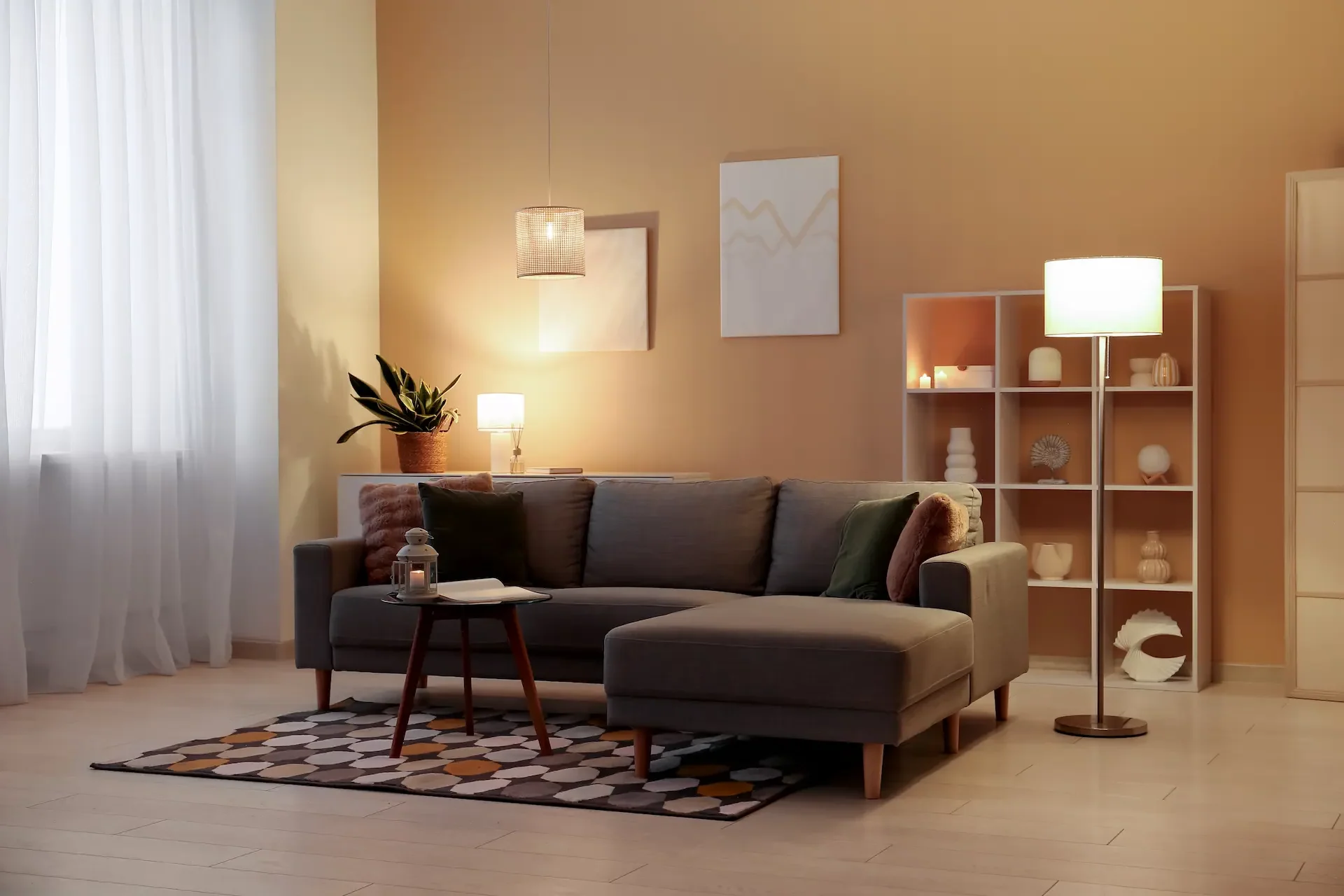 Un salon chaleureux avec un canapé d'angle gris, des coussins et un tapis à motifs. Un éclairage doux, provenant de lampadaires et de lampes de table, crée une ambiance accueillante.