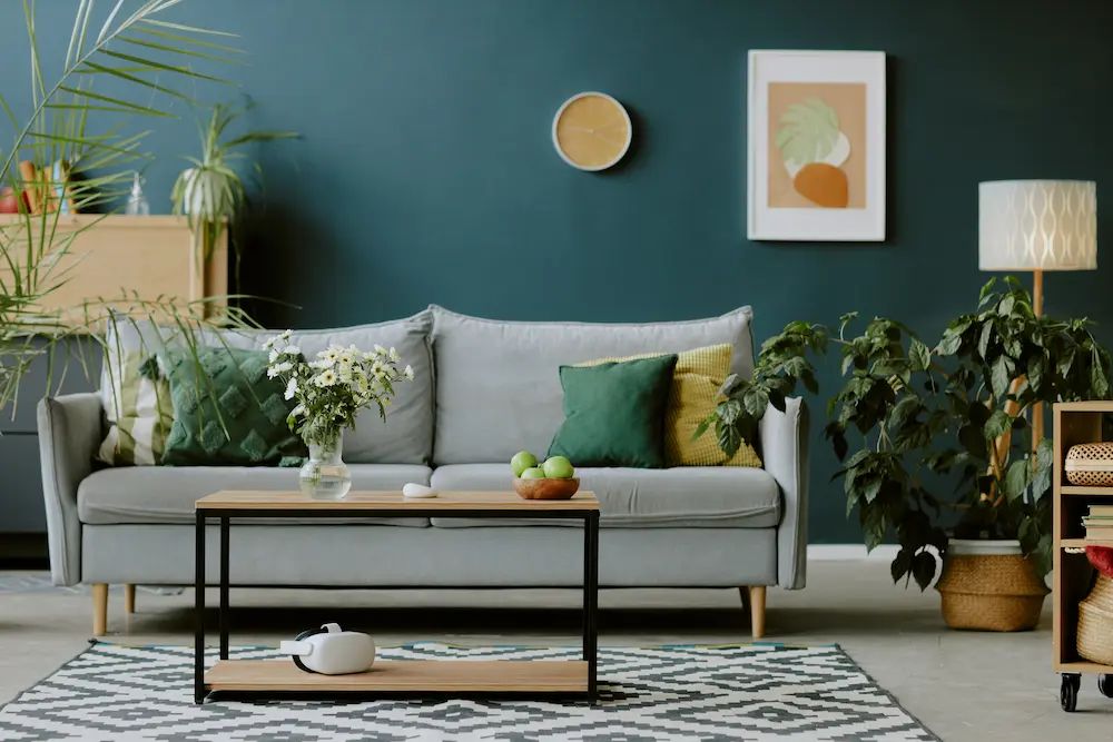 Salon élégant avec un canapé gris orné de coussins verts et jaunes, et une table basse en bois agrémentée d'objets décoratifs. Des plantes et un lampadaire contribuent à créer une ambiance à la fois chaleureuse et moderne.