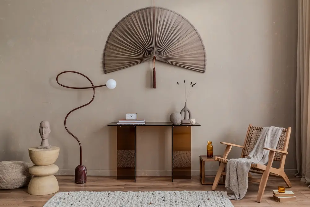 Un intérieur minimaliste aux tons beiges présente une décoration murale en forme d'éventail tressé, une console moderne, des sculptures artistiques et un fauteuil en bois confortable recouvert d'une couverture.