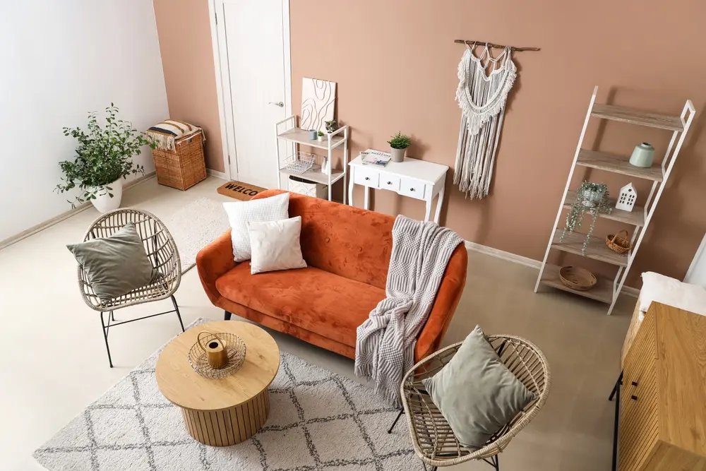 Salon chaleureux avec un canapé couleur terre cuite, des coussins blancs et un plaid gris. La décoration rustique comprend des fauteuils en osier, une table ronde en bois et des plantes.