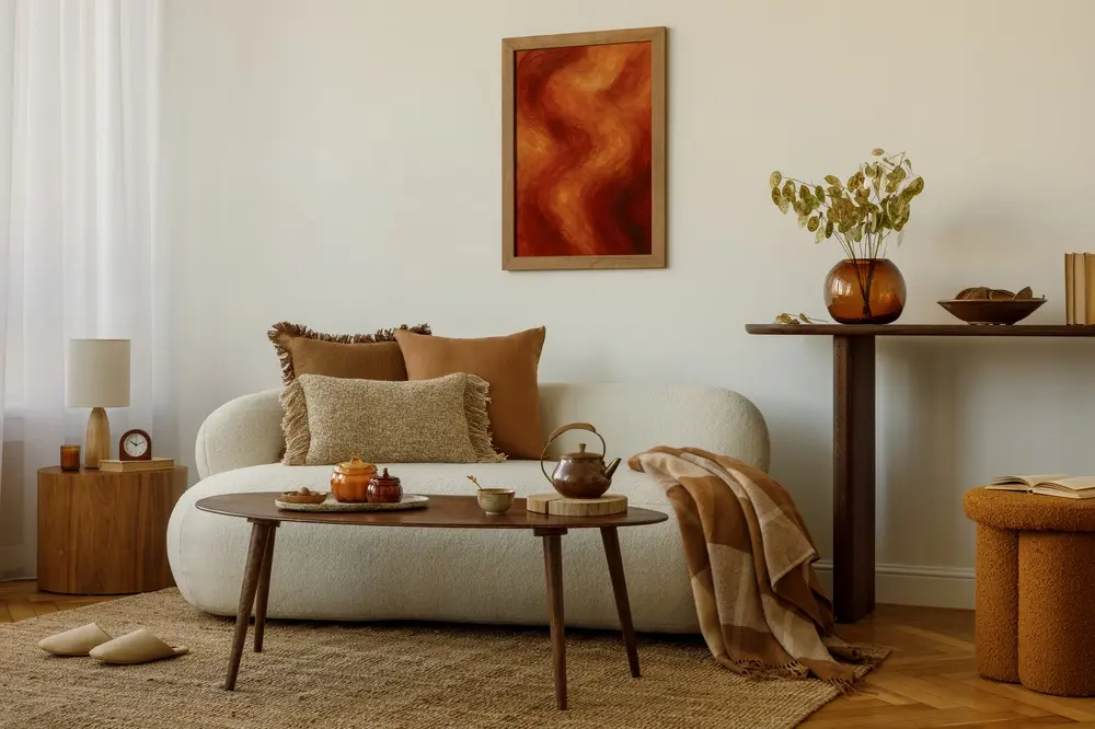 Salon chaleureux avec un canapé crème, des coussins marron et une table basse en bois. Les tons chauds dominent la décoration, rehaussée par un tableau abstrait rouge au mur.