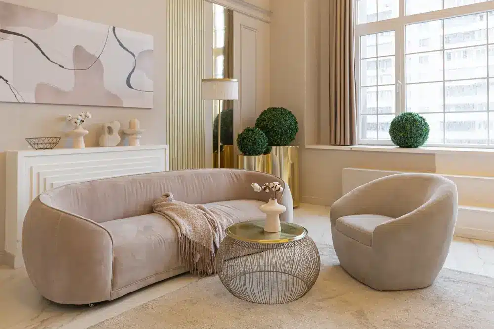 Salon élégant avec canapé et fauteuil beiges, table basse ronde en fil de fer, œuvres d'art abstraites et touches dorées. Grande fenêtre laissant entrer une douce lumière naturelle.