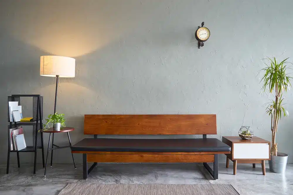 Salon cosy au décor moderne : banquette en bois, lampadaire, petites tables d’appoint, horloge et plantes. Ambiance chaleureuse et accueillante, dans un style minimaliste.