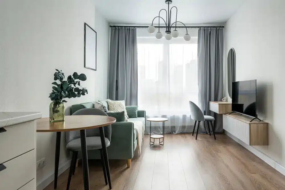 Salon moderne avec un canapé vert, un parquet et une table ronde agrémentée de plantes. Grande fenêtre aux rideaux gris, sous un élégant lustre.