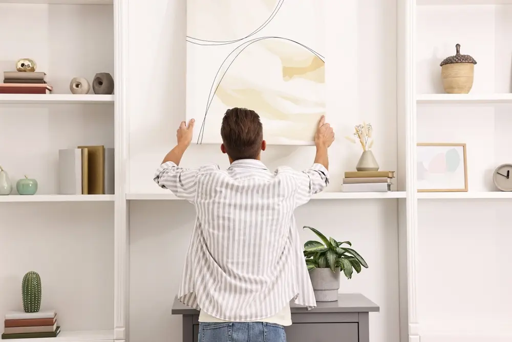 Une personne en chemise à rayures accroche des œuvres d'art abstrait au mur, entre des étagères blanches. La pièce, soigneusement décorée de livres, de plantes et de poteries, offre une atmosphère calme et élégante.